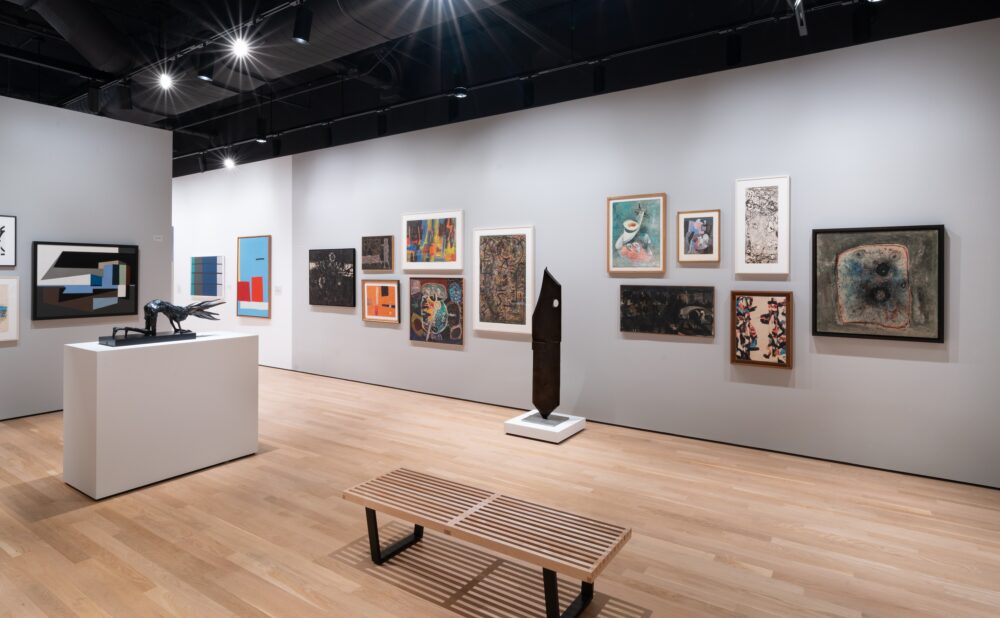Art gallery with various paintings on gray walls and a black sculpture on a pedestal; a wooden bench is in the foreground.