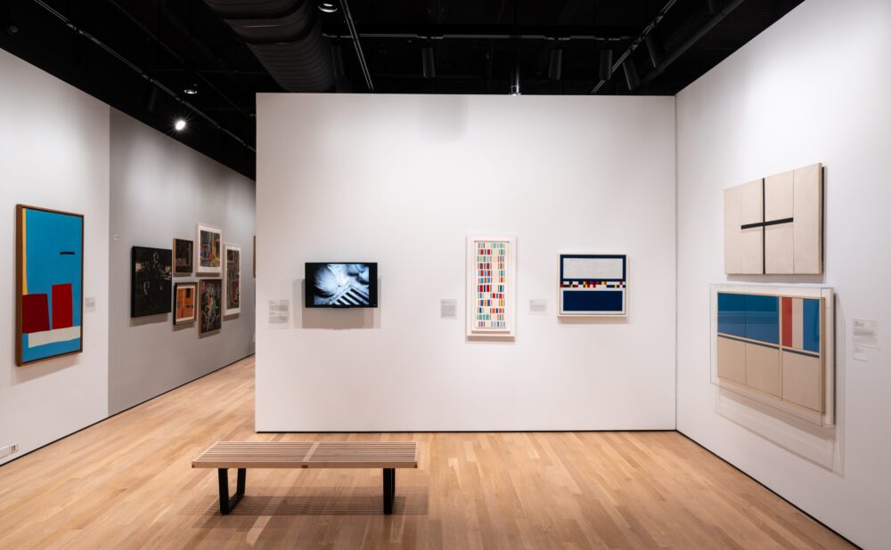 Art gallery with various paintings, a digital screen, and a wooden bench on a light wooden floor.