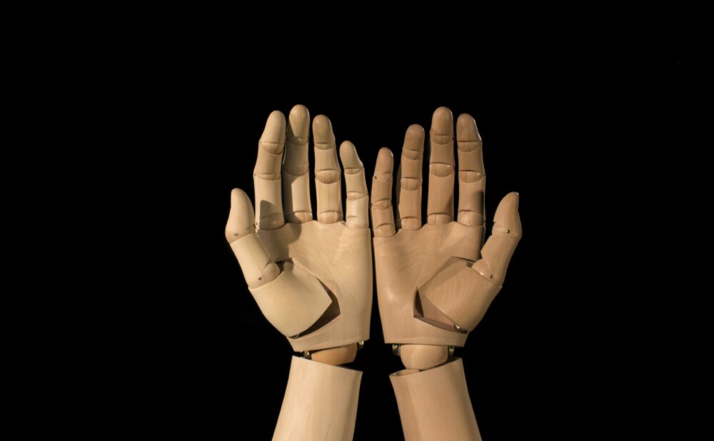 A pair of puppet wooden hands aside each other, palms up, facing the viewer, against a black screen
