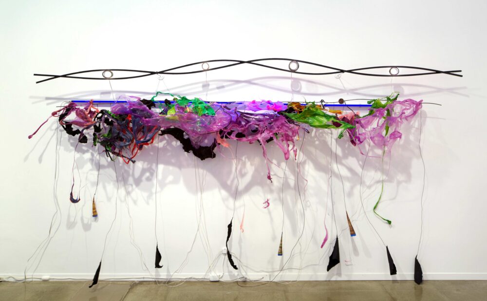 A sculpture comprising of a steel rod twisted in the shape of a DNA strand, hanging above a jubilant array of melted green, pink, and purple plastics and florescent lights. Tendrils of plastics reach down to the gallery floor.