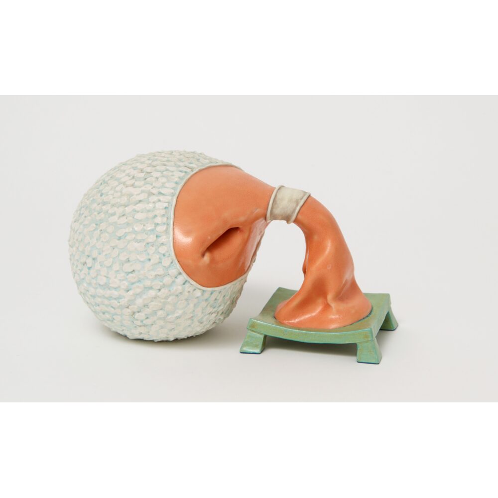 A ceramic sculpture in white, orange, and green takes on anthropomorphic qualities, its textured white head drooping down to the floor alongside its skinny orange neck.