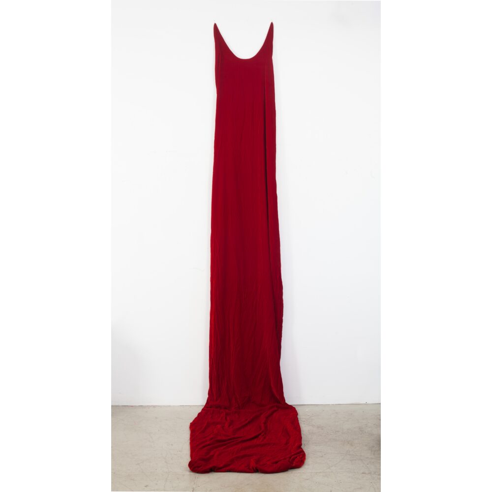 A sculpture of deep red velvet fabric takes the form of an absurdly long sleeveless dress; the shoulders of the dress are high up on the wall, and the body of the form cascades down the wall and pools onto the gallery floor.