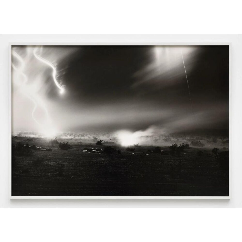 A black-and-white photograph in a thin white frame shows a vast nighttime, desert landscape on which military vehicles ride and conduct training