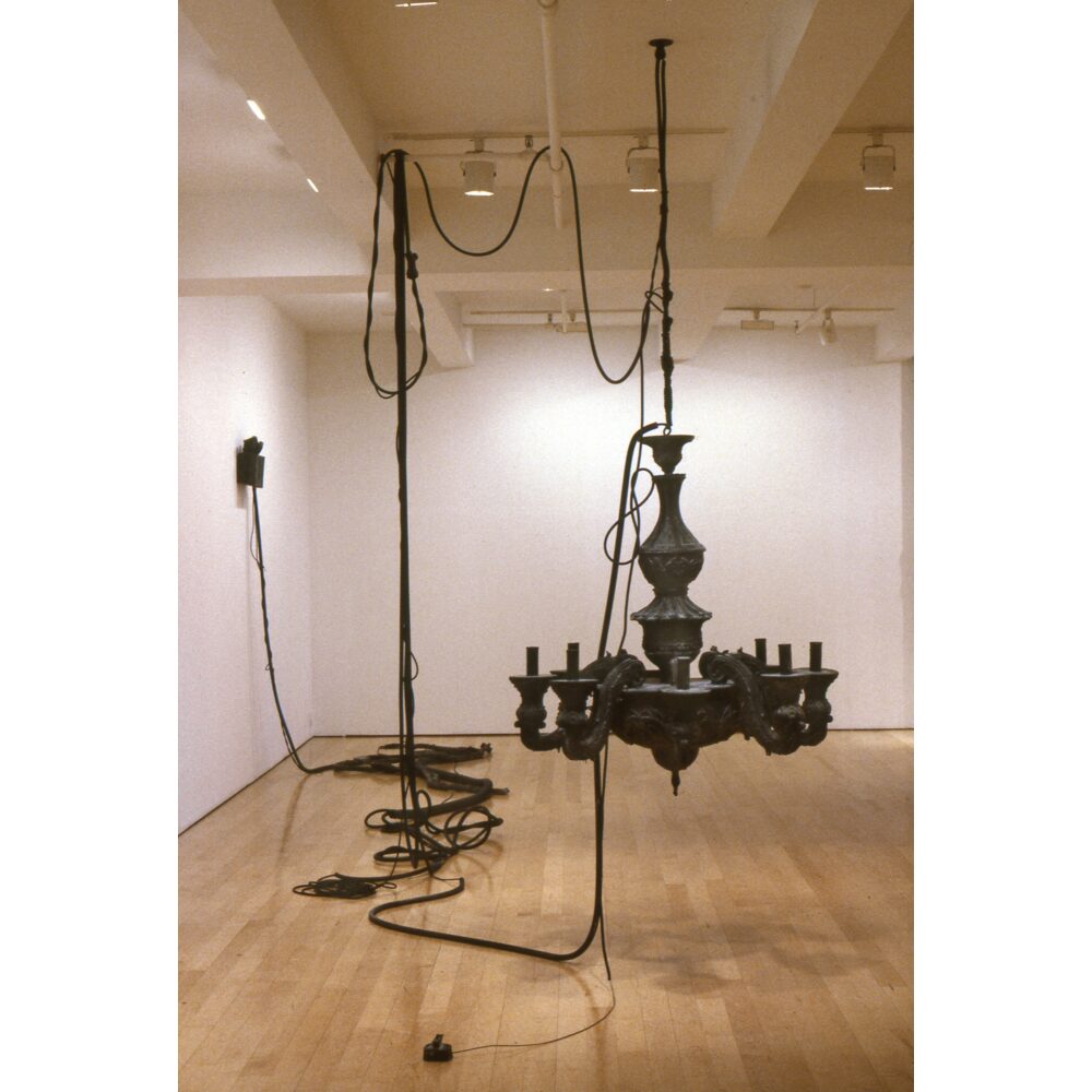 A black rubber installation fills a gallery, with a large suspended chandelier form as the central component, surrounded by carefully suspended and looped cords and faux electrical components, including an EXIT sign