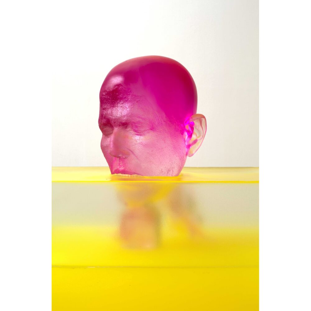 A sculpture comprising of a woman's head cast in magenta and transparent resin, sunk partially into a block of yellow and transparent resin.