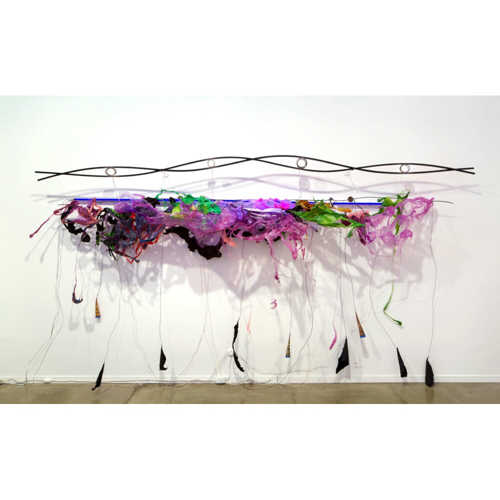 A sculpture comprising of a steel rod twisted in the shape of a DNA strand, hanging above a jubilant array of melted green, pink, and purple plastics and florescent lights. Tendrils of plastics reach down to the gallery floor.