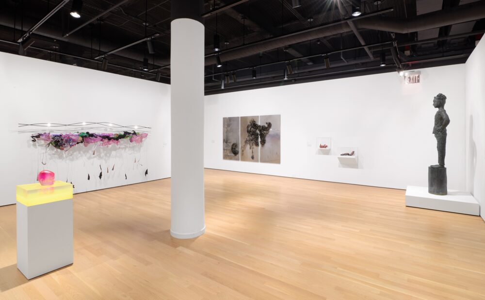 Modern art museum with resin sculpture on a pedestal, colorful wall sculpture with lights, a bronze figurative sculpture, and multimedia artwork with paper and reflective surfaces.