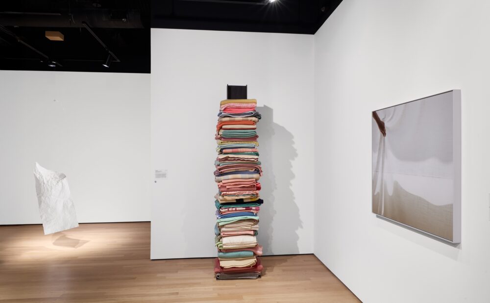 Modern art museum showing one paper sculpture hanging from the ceiling, one textile sculpture standing against the wall, and a photograph mounted to the wall