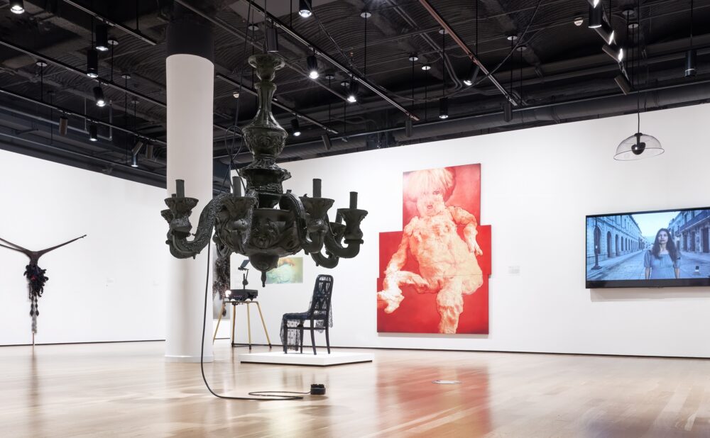 Modern art museum featuring a projector, a painting on the wall, a video on a monitor, a lace-covered chair on a platform, a textile sculpture hanging on the wall, and a chandelier-like black resin sculpture hanging from the ceiling.
