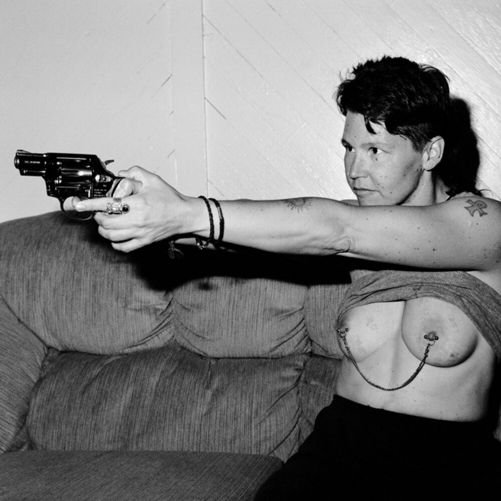 Black-and-white portrait of a person sitting indoors holding a handgun toward the camera frame. Their breasts, bearing a nipple chain, are exposed. (Alt-text generated by AI)