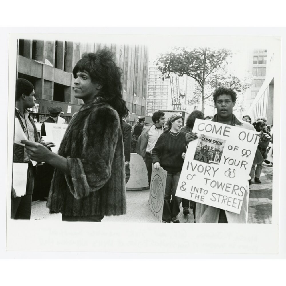 Black-and-white photo showing individuals outdoors holding protest signs in an urban setting. (Alt-text generated by AI)
