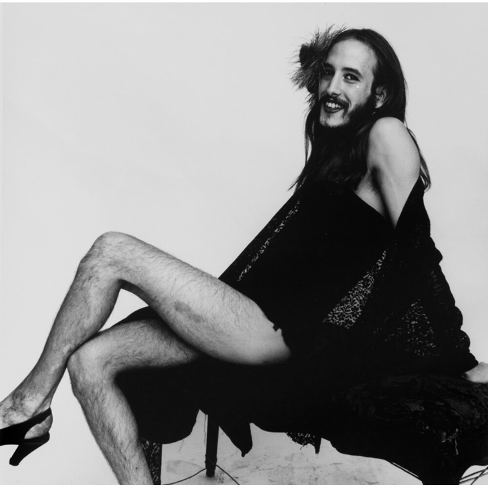 Black-and-white portrait of a long-haired, smiling, bearded person sitting in a dynamic pose against a neutral background. (Alt-text generated by AI)