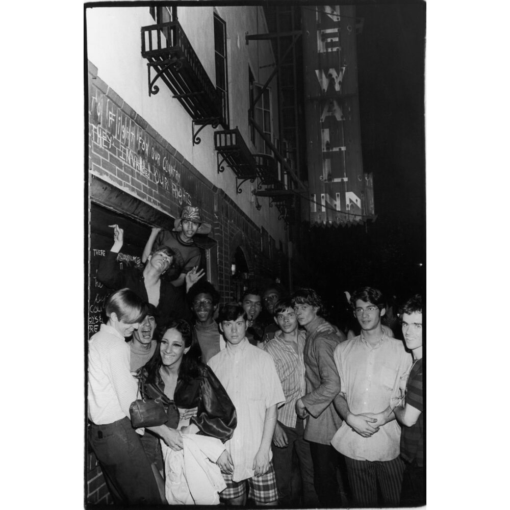 Vertical black-and-white photograph showing a crowd gathering outdoors beneath street signs and buildings. (Alt-text generated by AI)
