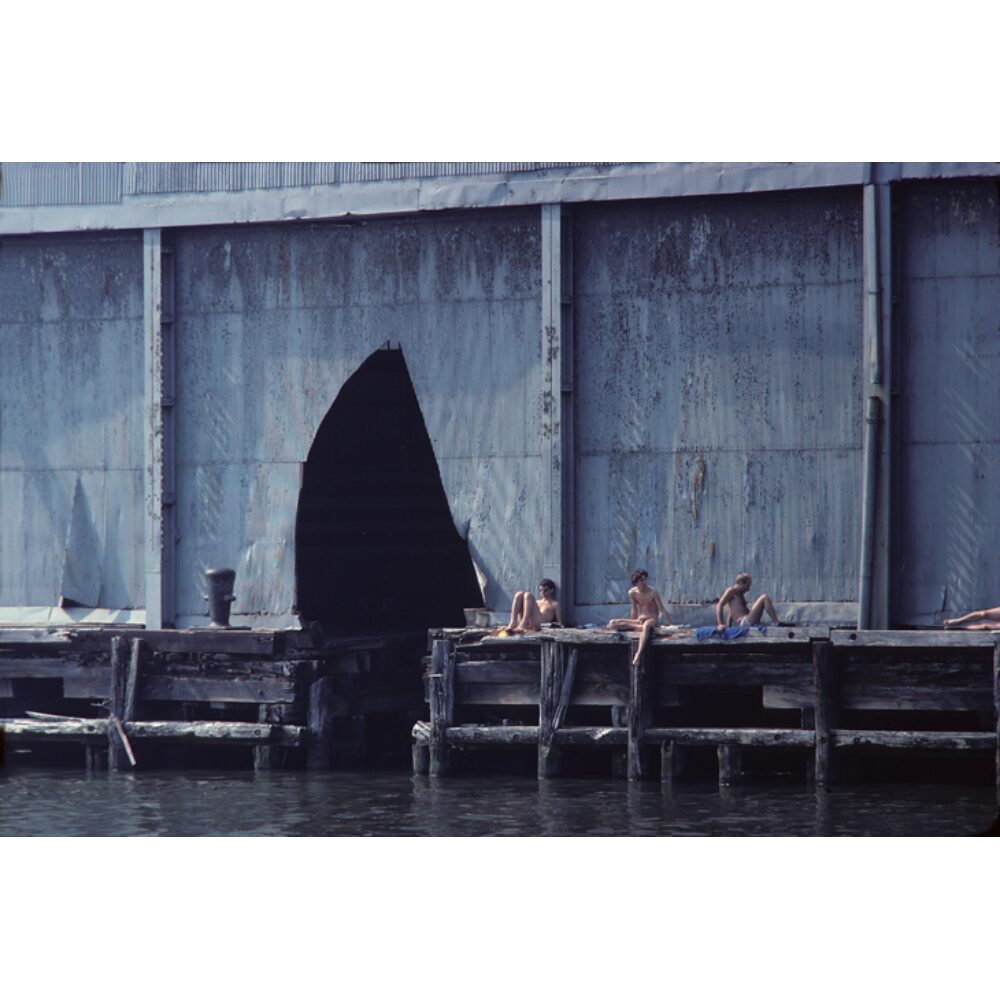 Black-and-white photograph depicting figures sitting outdoors on a rundown dock. (Alt-text generated by AI)