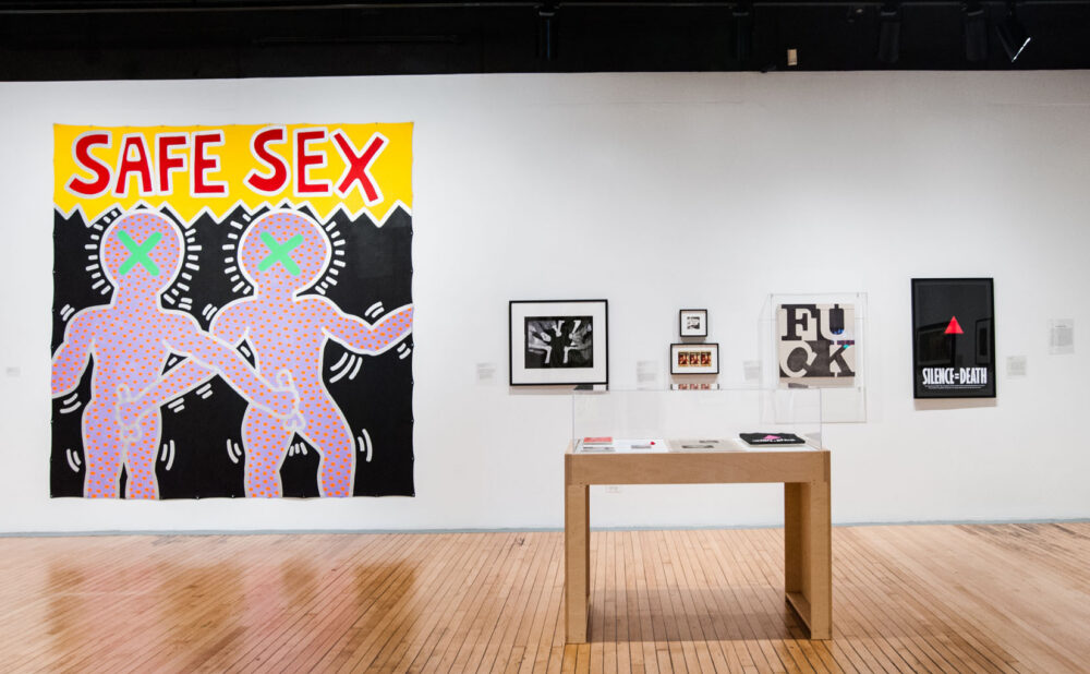 Gallery with a large poster reading "SAFE SEX" mounted above a wooden display table surrounded by smaller framed prints. (Alt-text generated by AI)