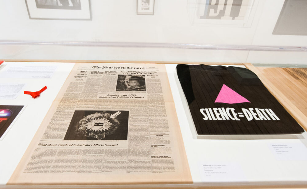 Close-up top-down view of printed archival materials, newspapers, and a black t-shirt with the text "SILENCE=DEATH" laid flat in a display case. (Alt-text generated by AI)