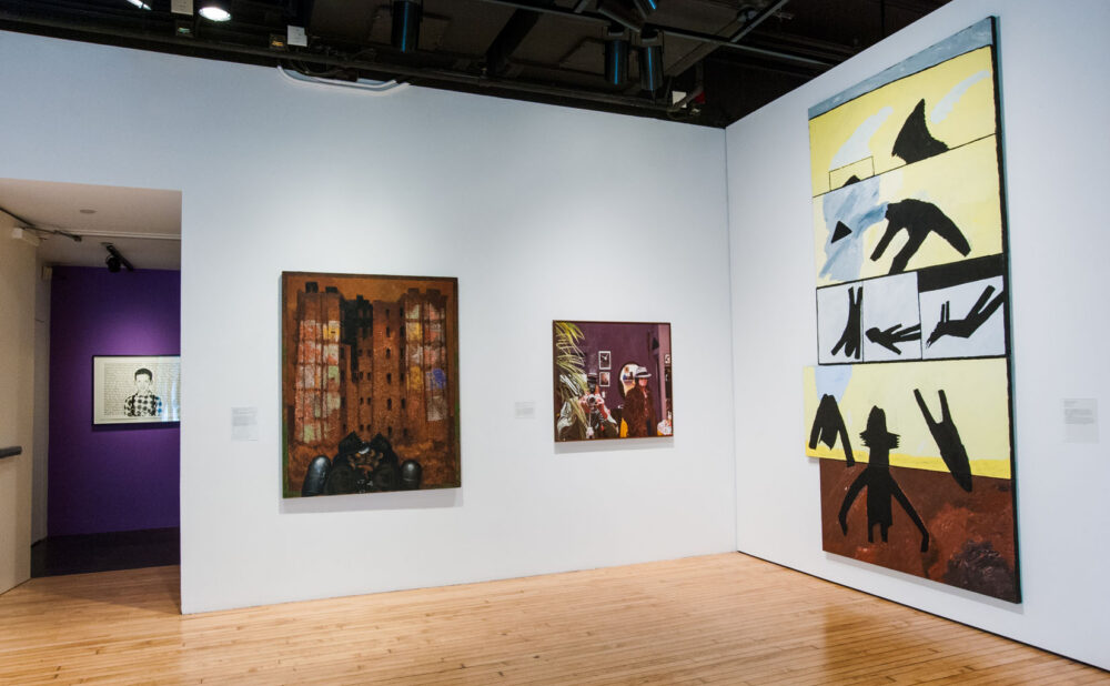 High-ceilinged gallery featuring a tall, vertical graphic artwork alongside several smaller framed wall works. A down stairwell with purple interior is visible on the left. (Alt-text generated by AI)