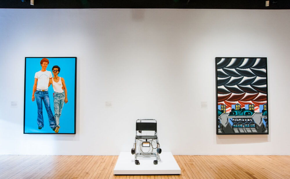 Gallery showing a portrait with a blue background on the left wall, a central sculpture on a white pedestal, and a geometric pattern artwork on the right. (Alt-text generated by AI)