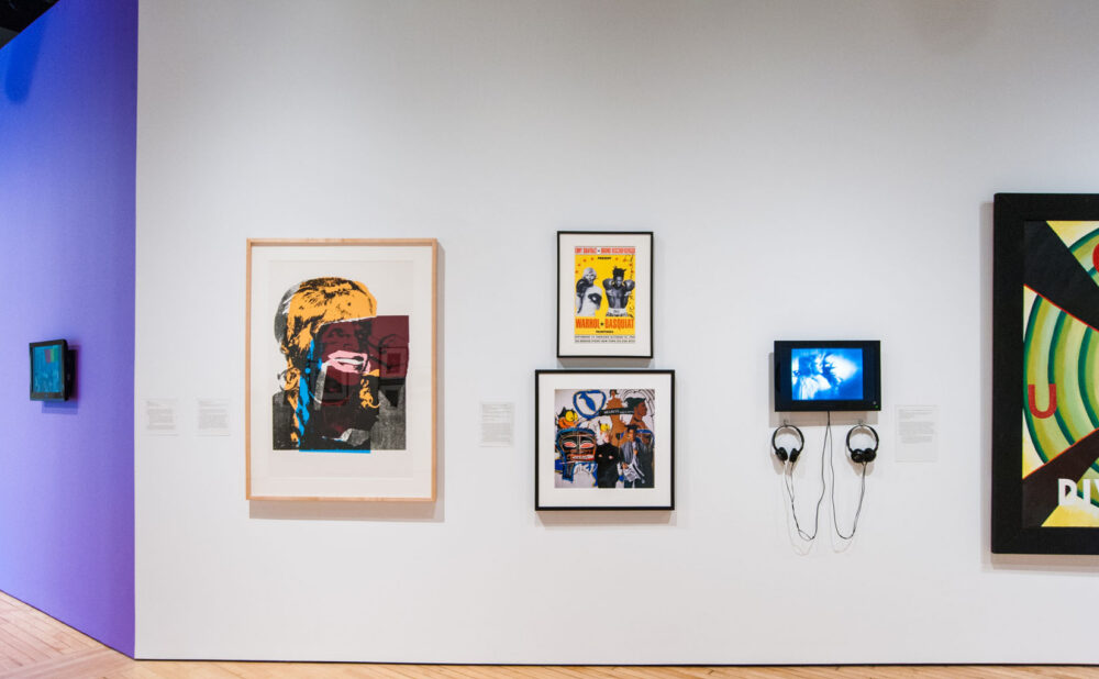 Gallery wall featuring several framed prints, a small video display monitor, and a purple accent wall on the left. (Alt-text generated by AI)