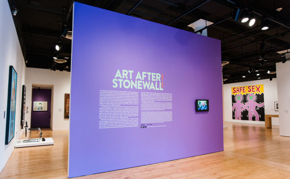 Entrance view of the exhibition featuring a large bright purple freestanding wall with title text. (Alt-text generated by AI)