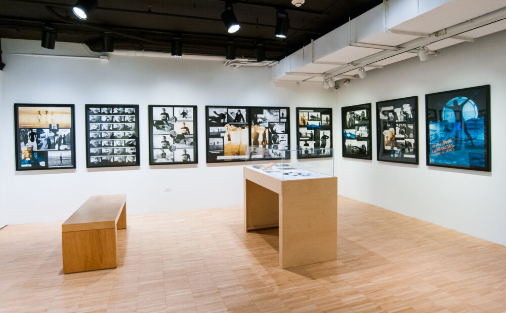Gallery room with a row of framed artworks along the wall and a bench and display table in the center. (Alt-text generated by AI)