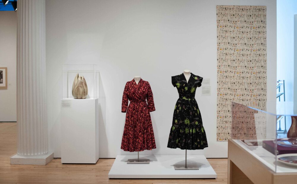 Exhibition display showing two mannequin forms dressed in patterned mid-century dresses alongside white pedestals and wall hangings. (Alt-text generated by AI)