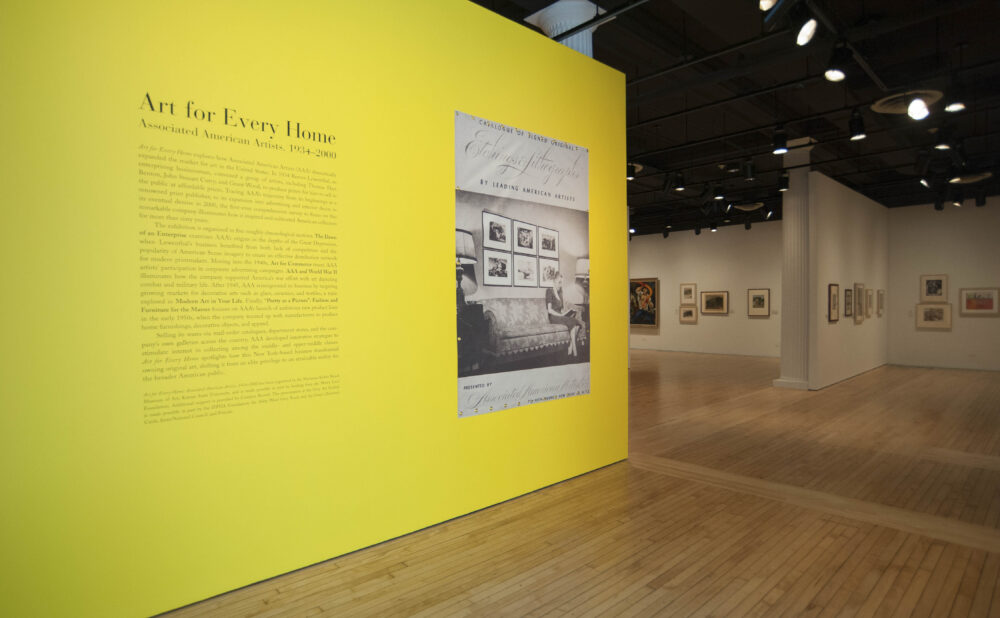 Perspective view of the exhibition entry featuring a bright yellow accent wall printed with introductory text and wall graphics. (Alt-text generated by AI)