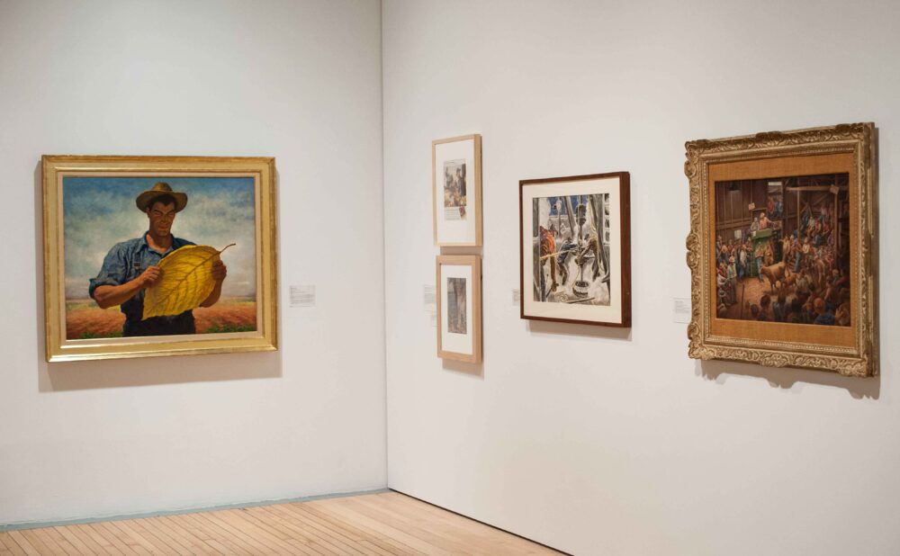 Corner gallery view displaying framed regionalist paintings and prints mounted on adjacent white walls. (Alt-text generated by AI)