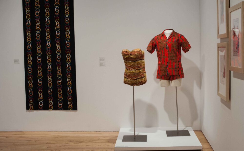Exhibition platform featuring a red patterned shirt on a dress form, a strapless women's bathing suit on another dress form beside it, and a patterned textile hanging on the wall behind. (Alt-text generated by AI)