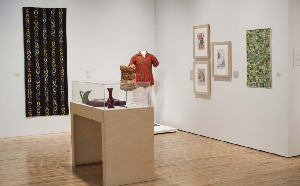 Gallery section featuring a central wooden display table in front of wall-mounted textile samples and framed artworks. (Alt-text generated by AI)