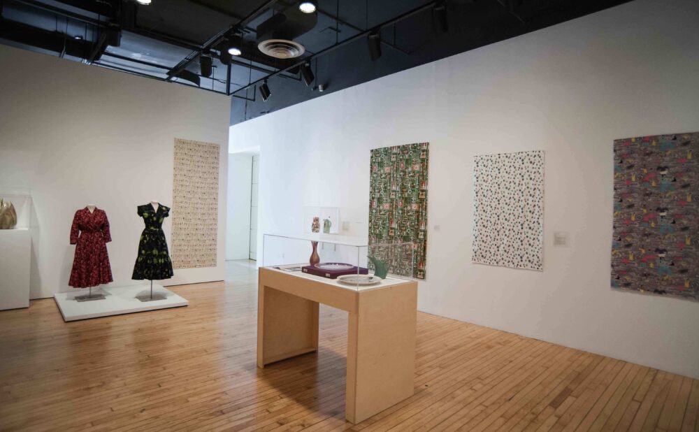 Wide exhibition view showing clothing displays, central vitrines, and patterned textile lengths hung on white gallery walls. (Alt-text generated by AI)