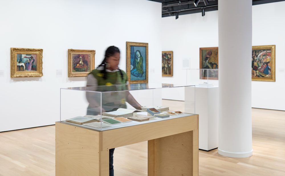 Modern art gallery with white walls and a column, with framed turn-of-the-century European paintings on the wall; at center, a Black woman examines items in a display case.