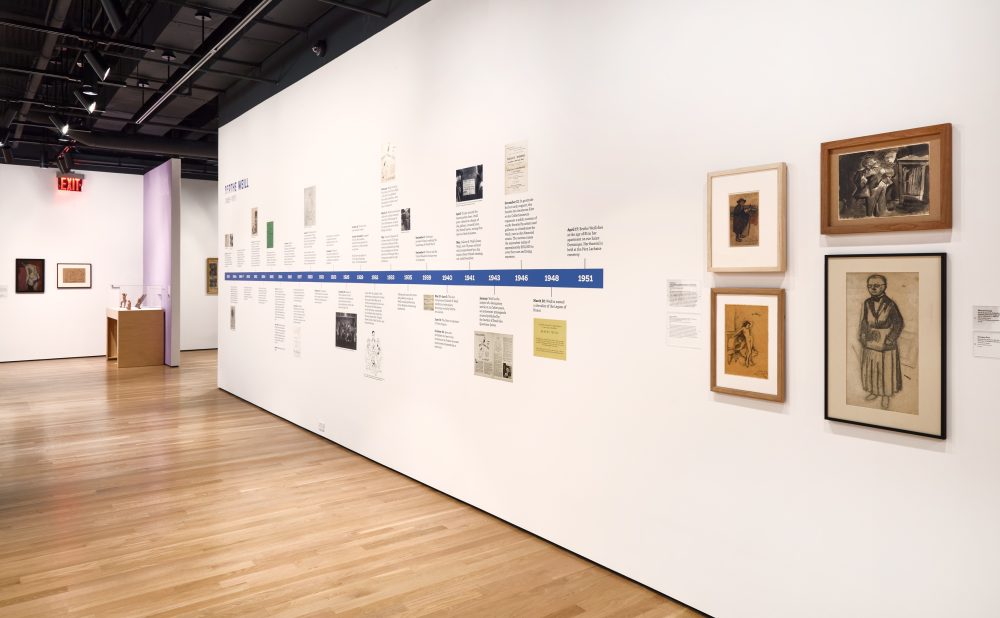 Art gallery with white walls, wooden floors, framed artworks, and a timeline.