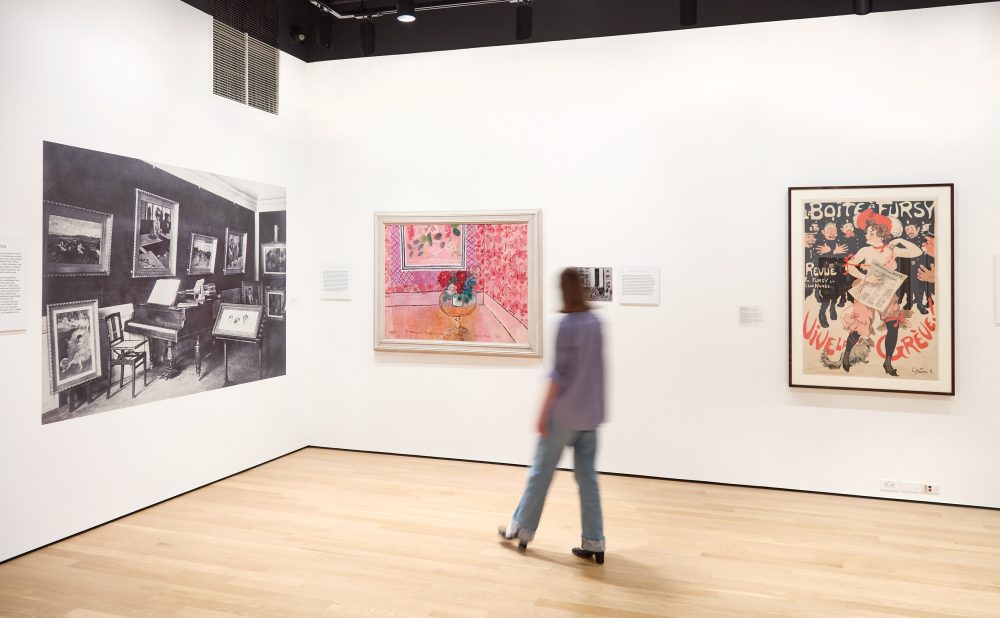 Art gallery with a mural, painting, and vintage poster, with a person walking through.