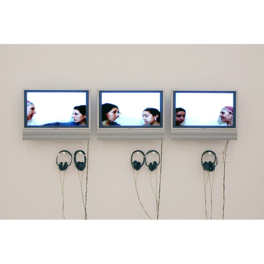 Video installation featuring three wall-mounted screens displaying video interviews with attached headphones. (Alt-text generated by AI)
