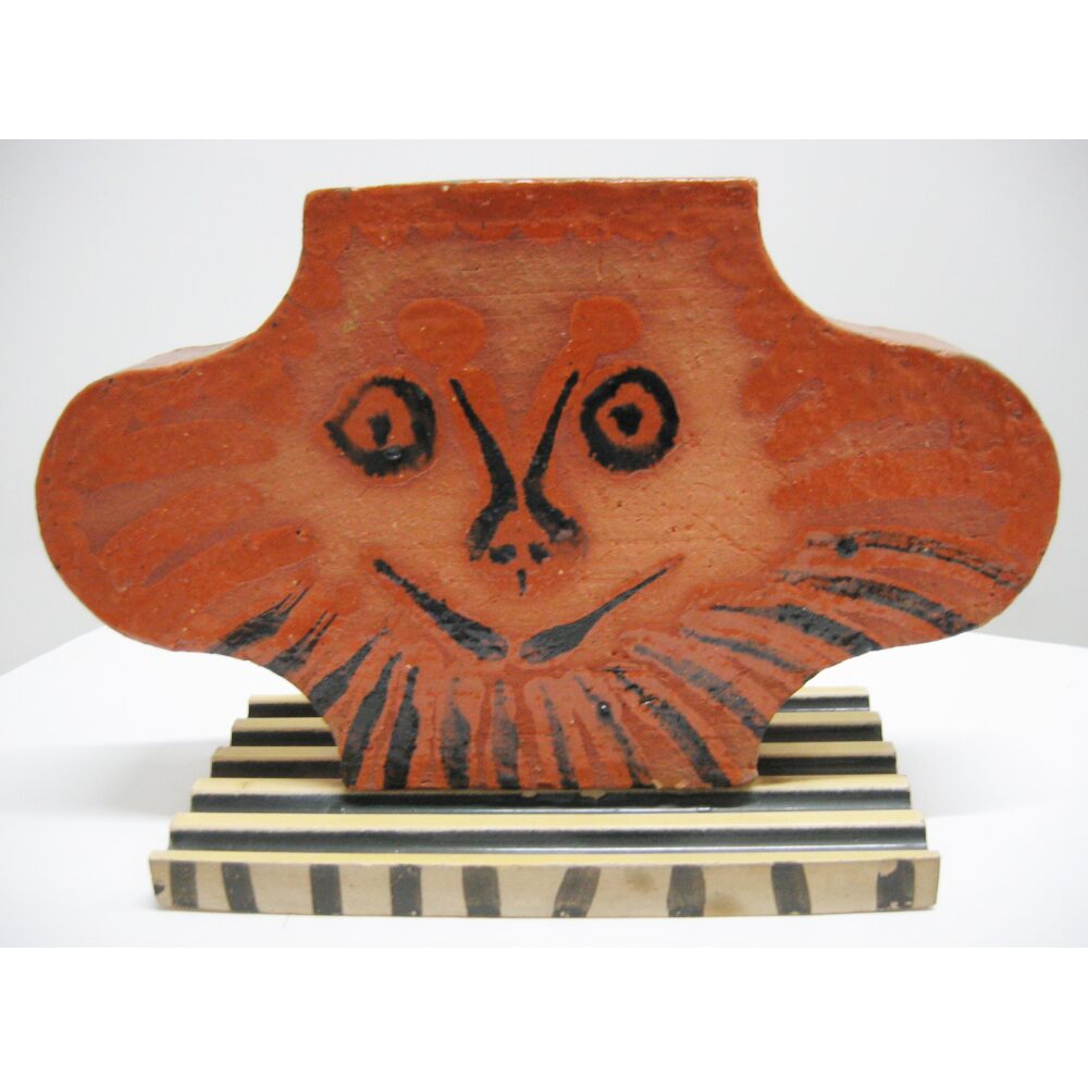 Terra cotta-red ceramic sculpture depicting a double-faced head on a striped pedestal base. (Alt-text generated by AI)