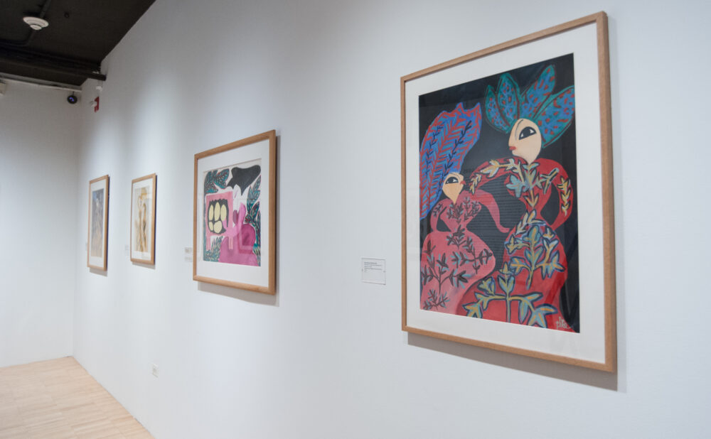 Perspective view along a white gallery wall featuring framed colorful paintings, including a large artwork on the right. (Alt-text generated by AI)