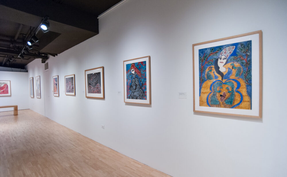 Angled perspective along a long gallery wall lined with framed, brightly colored paintings above polished wood flooring. (Alt-text generated by AI)