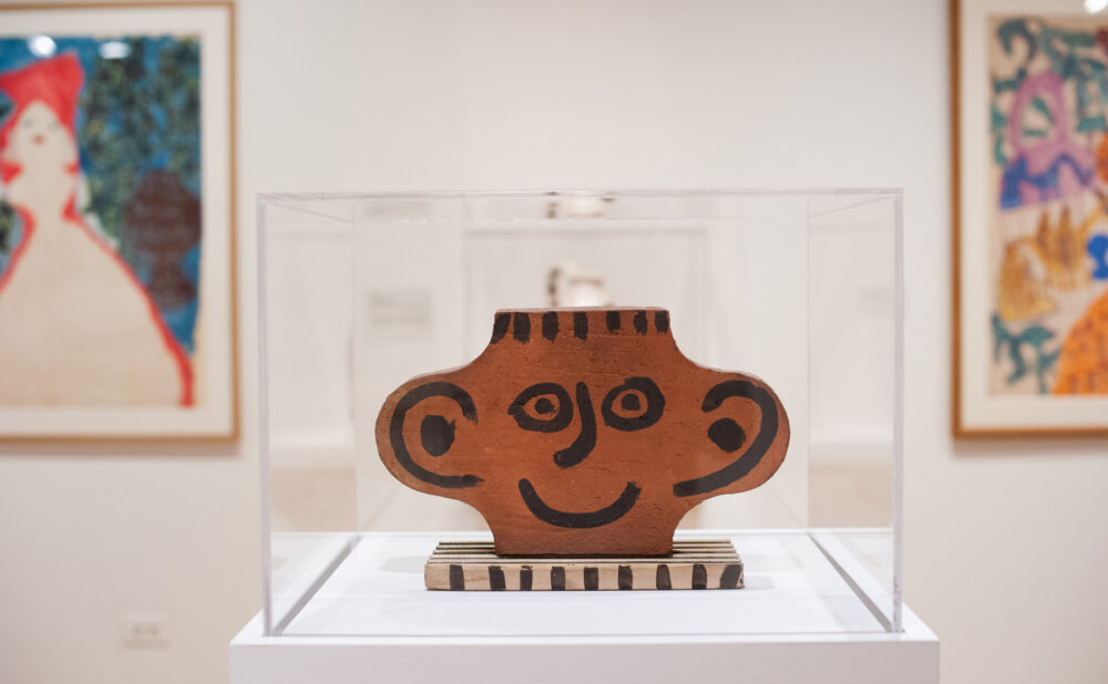 Close-up view of a ceramic sculpture featuring a whimsical face enclosed in a clear display case, flanked by framed paintings in the background. (Alt-text generated by AI)