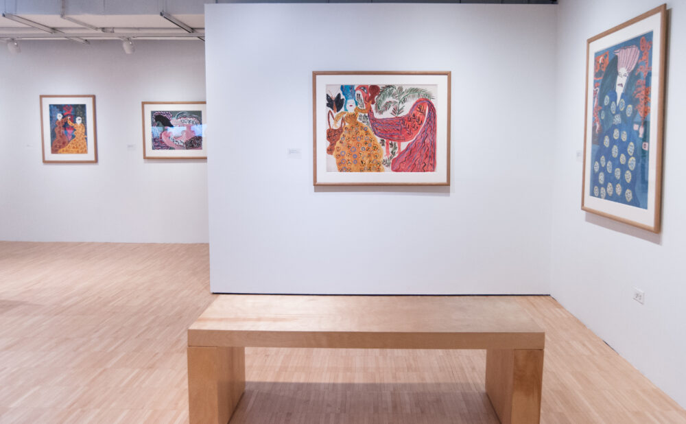 Open gallery space with a wooden bench centered in front of a white wall mounted with framed paintings. (Alt-text generated by AI)