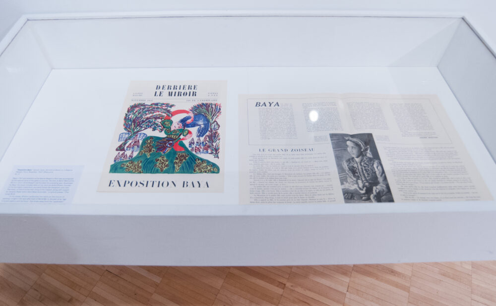 Vitrine display case containing opened exhibition catalogs and printed archival materials. (Alt-text generated by AI)