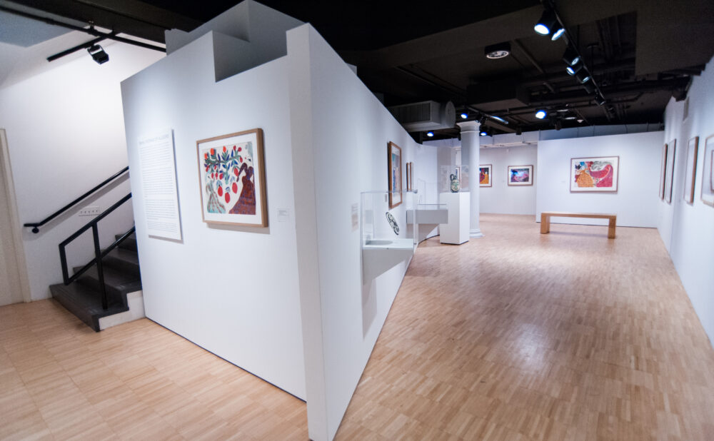 Wide architectural view of the gallery interior showing white exhibition partition walls, light wood flooring, and a staircase on the left. (Alt-text generated by AI)
