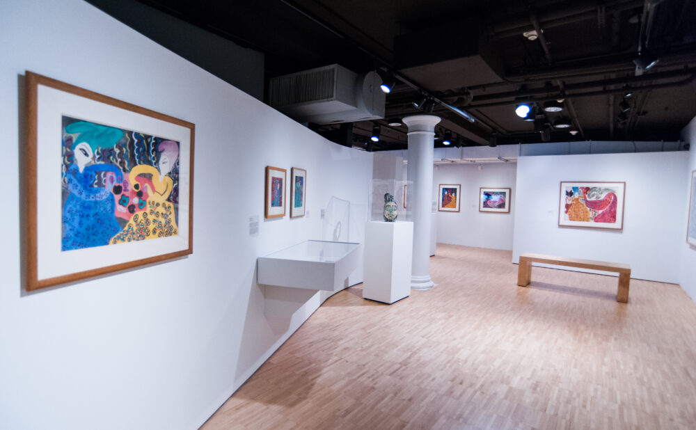 Gallery view looking down a central corridor with a large framed painting on the left foreground wall and additional works in the distance. (Alt-text generated by AI)