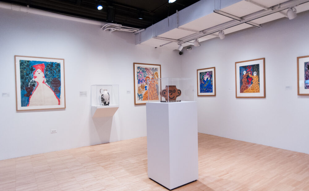 Gallery room featuring a central white pedestal displaying a small ceramic sculpture surrounded by framed colorful paintings on the walls. (Alt-text generated by AI)