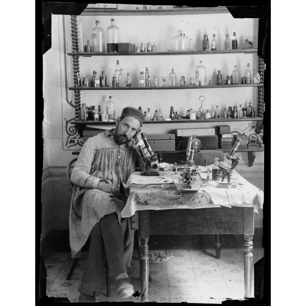 Black-and-white historical photograph of Santiago Ramón y Cajal sitting at a desk in his laboratory using a microscope. (Alt-text generated by AI)