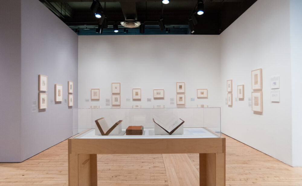 Wooden display case holding open books under glass cases in front of a white wall hung with small framed drawings. (Alt-text generated by AI)