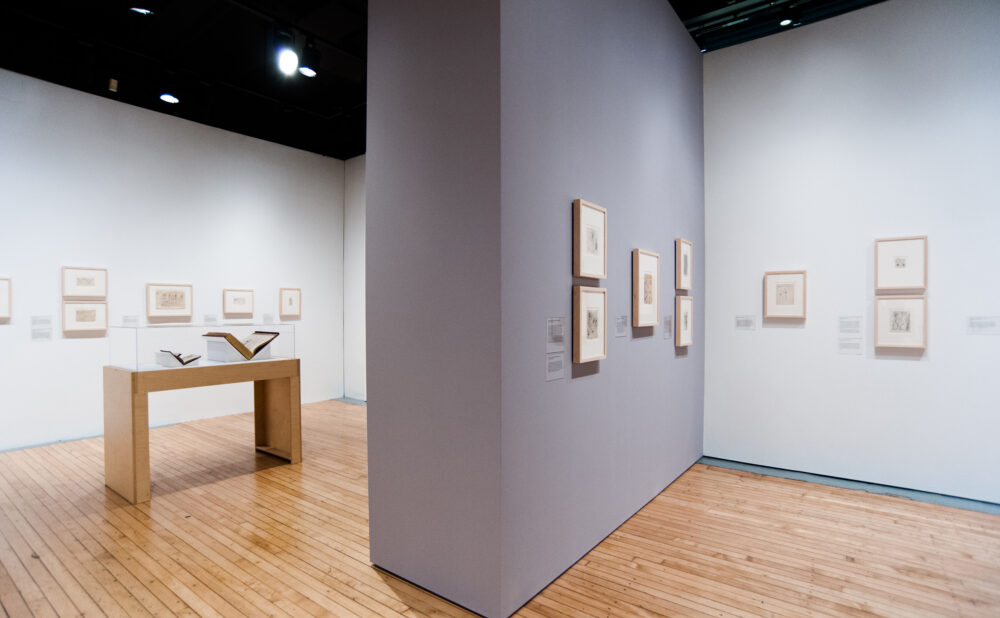 Wide view of a gallery room showcasing framed wall works and a central wooden display case. (Alt-text generated by AI)
