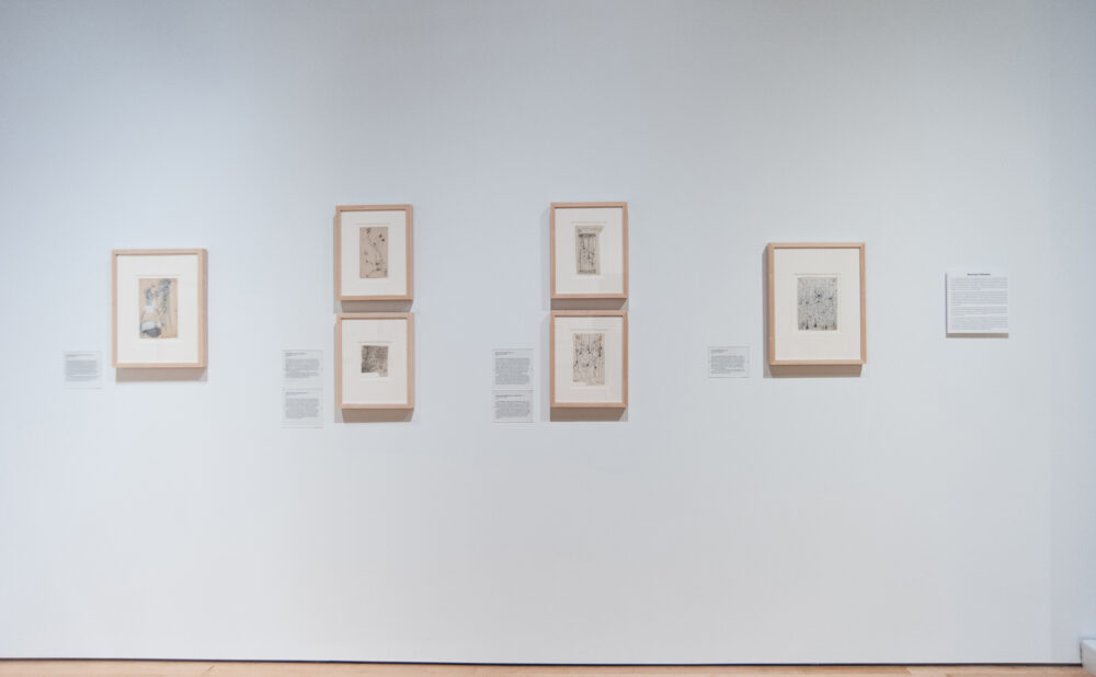 White gallery wall displaying five small framed artworks with wall labels arranged horizontally. (Alt-text generated by AI)