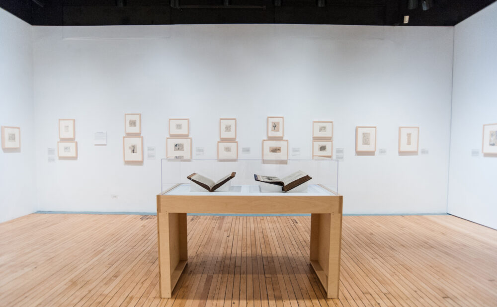 Symmetrical view of a wooden display case with open books centered against a long white gallery wall filled with small drawings. (Alt-text generated by AI)