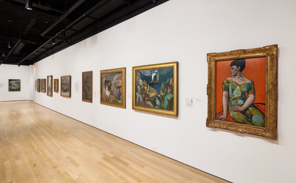 Art gallery with a row of framed paintings on white walls and a light wooden floor.